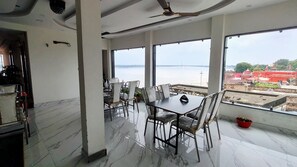 A dining area with a table and chairs, large windows with a view of a body of water and a bridge, and a ceiling fan. - Hotel Royal Shantii Inn (Varanasi)