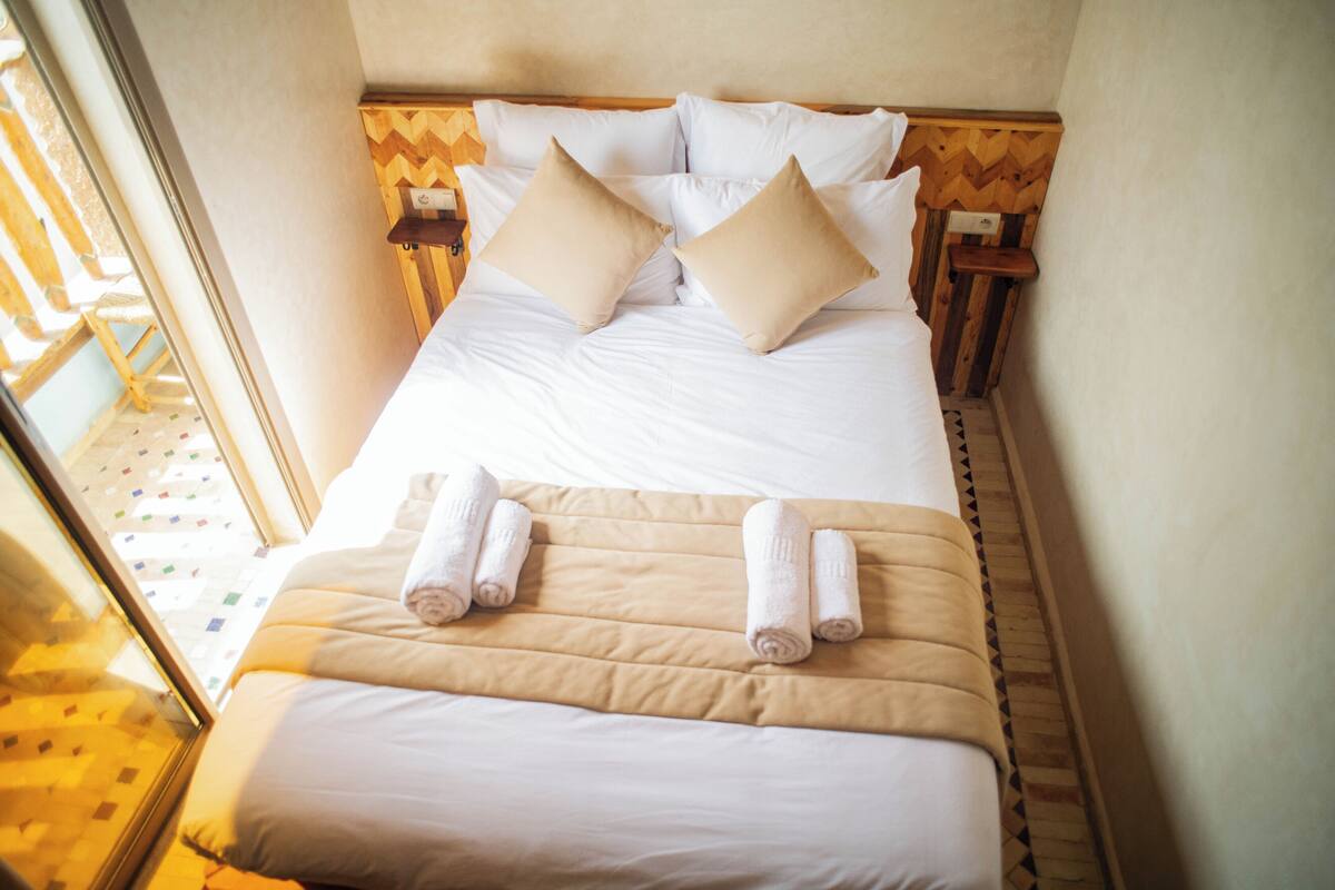 A neatly made bed with white linens and beige pillows, a wooden headboard, and two bedside tables with lamps.