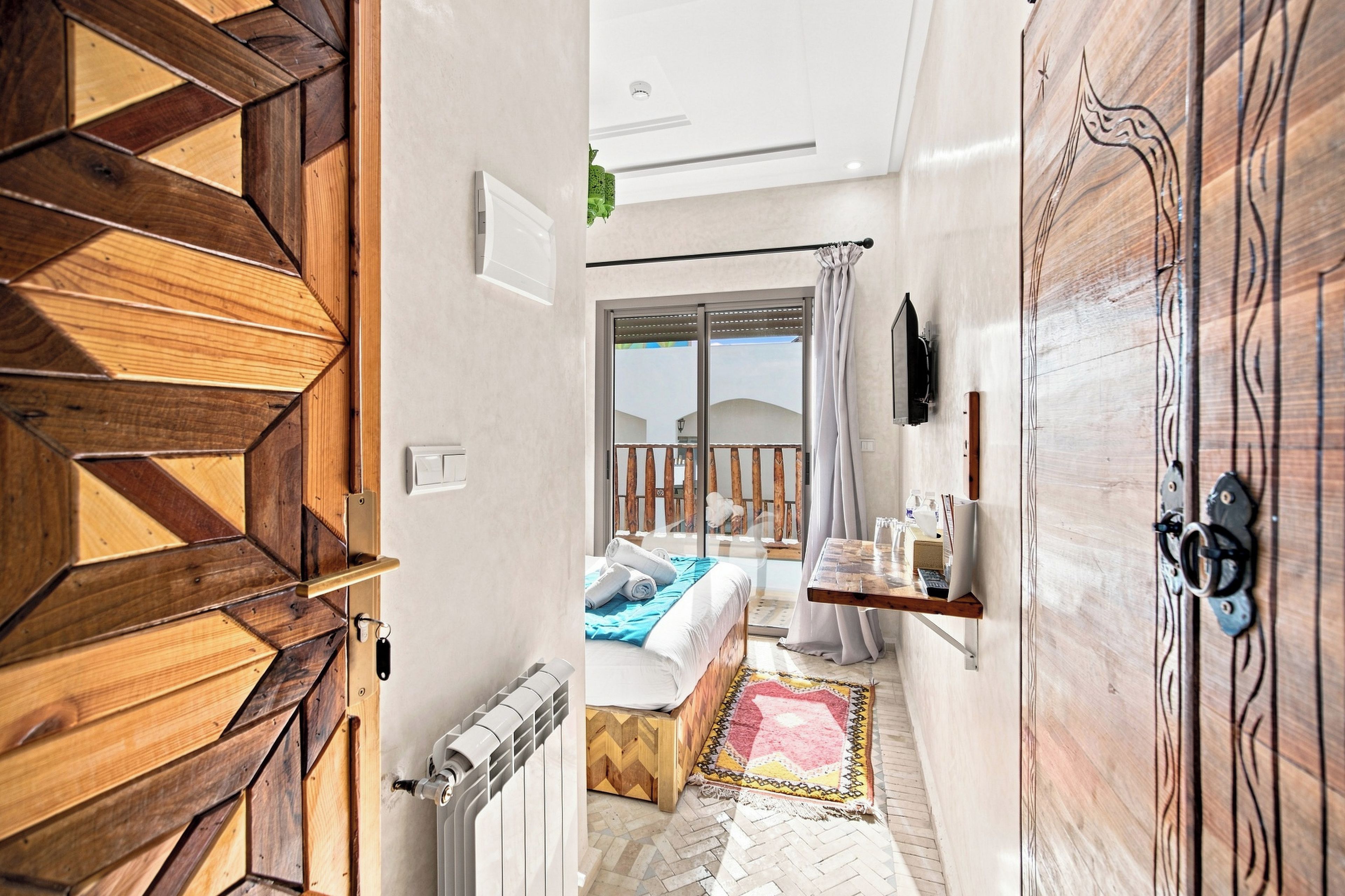 A hotel room with a wooden door, a bed with a blue blanket, a small table with a keyboard, and a view of the outside.
