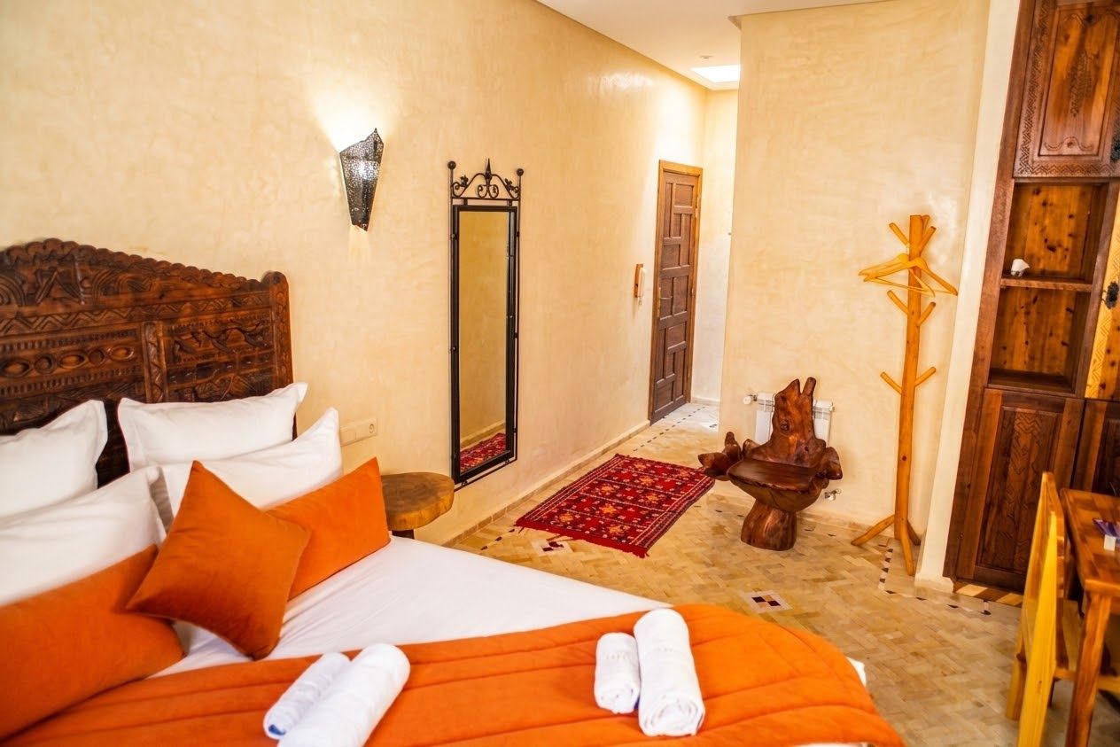A hotel room with a bed, a wooden headboard, a mirror, a wooden coat rack, and a small table with a decorative item.
