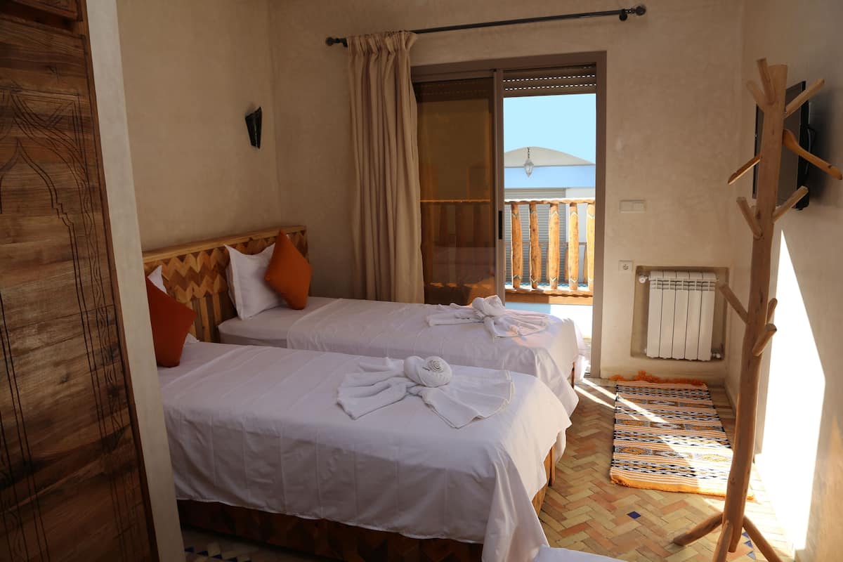A room with two beds, a wooden headboard, a window with a view, a wooden coat rack, and a patterned rug.