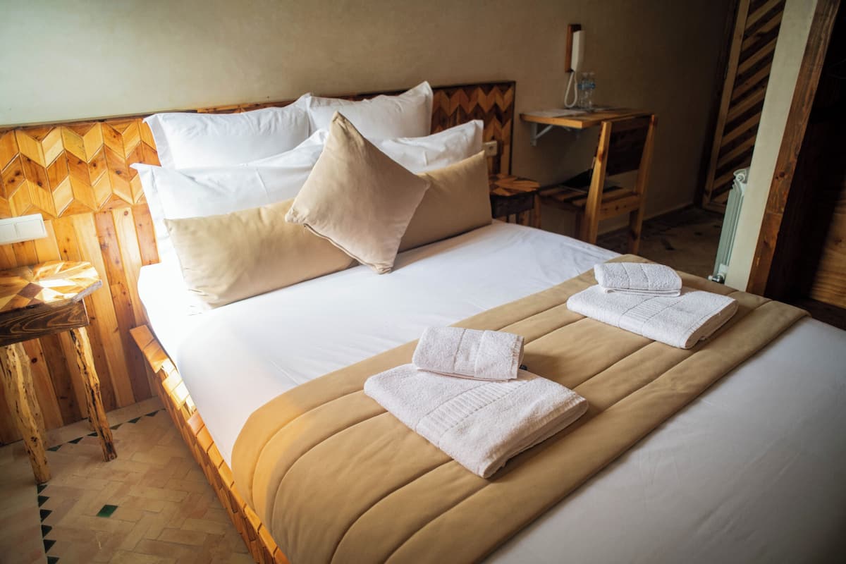 A neatly made bed with white linens and beige pillows, a wooden headboard, and a small wooden nightstand with a lamp.
