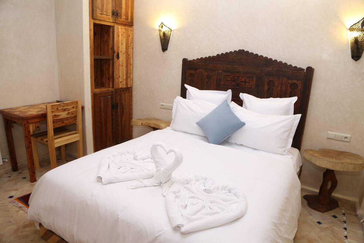 A neatly made bed with white linens and heart-shaped towels, a wooden headboard, and two wall-mounted light fixtures.