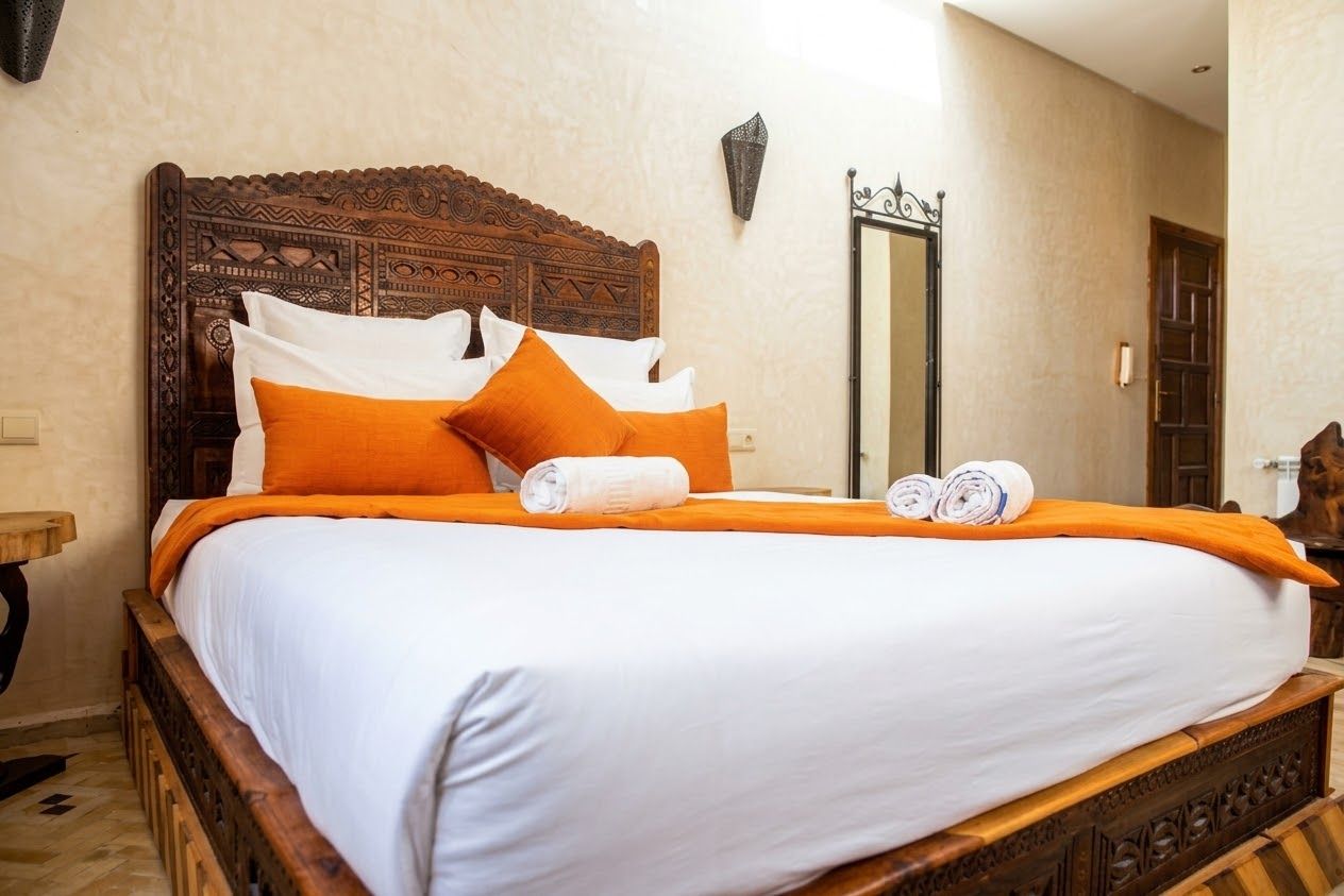A neatly made bed with a wooden headboard, white and orange bedding, and rolled towels.