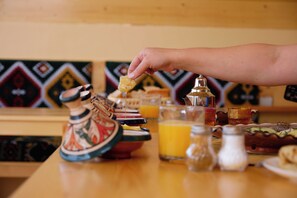 A hand holding a piece of food, with a table setting that includes a jug, glasses, and a plate with food. - Imsouane WOOD HOUSE (Imsouane)
