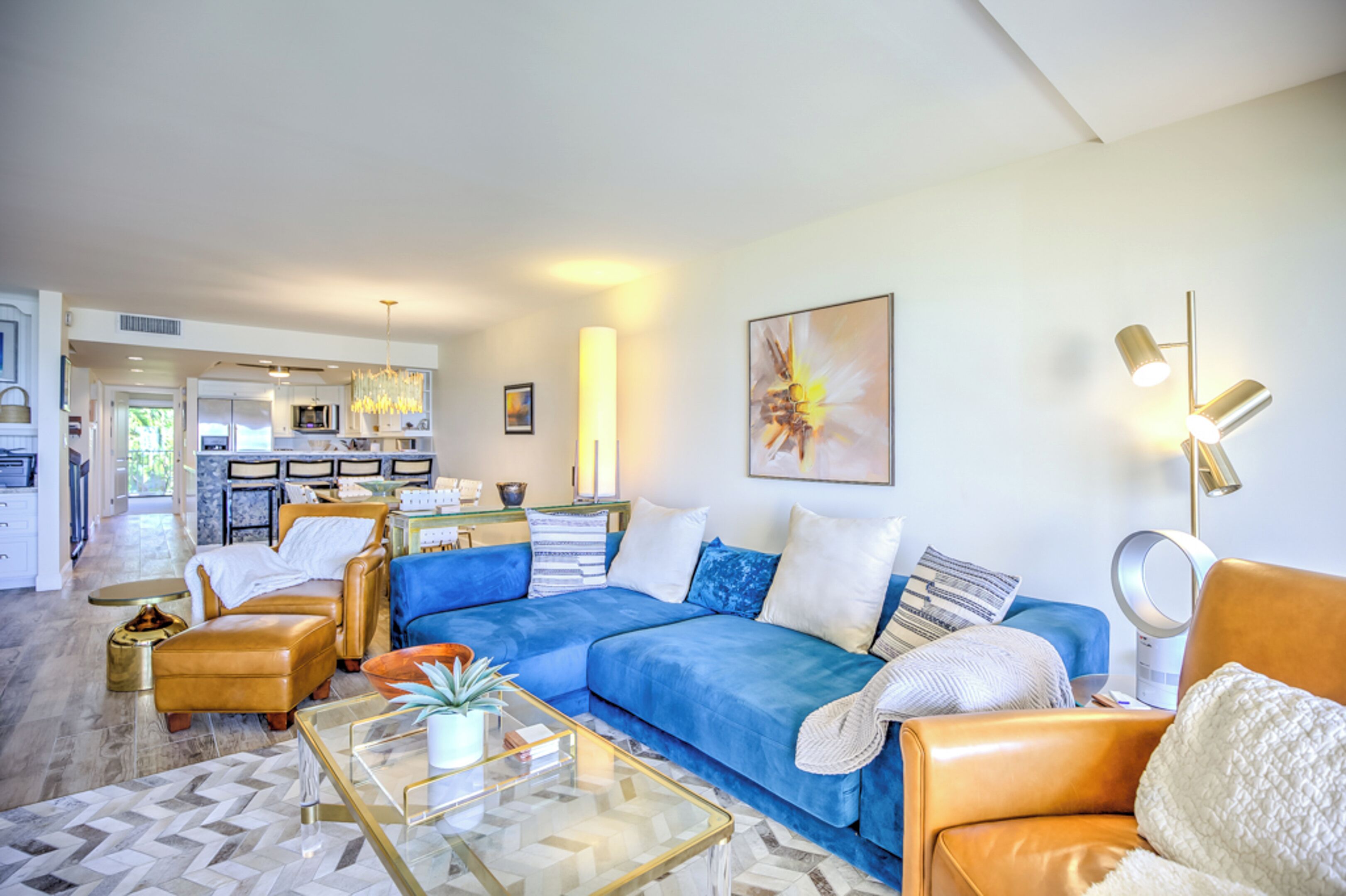 A modern living room with a blue sofa, leather armchairs, a glass coffee table, and a kitchen in the background.