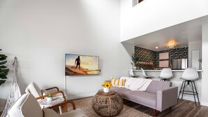 Una sala de estar moderna con un sofá gris, sillas blancas, una mesa de centro con un jarrón de girasoles y un televisor grande mostrando a un surfista.