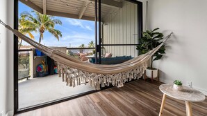 Una hamaca en una habitación con una puerta corredera de vidrio que da a un balcón con mobiliario exterior y palmeras.