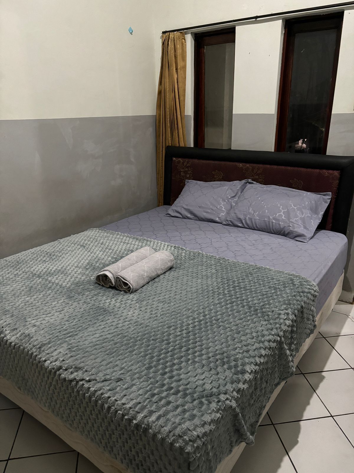 A neatly made bed with a grey blanket and a rolled towel, a window with curtains, and a tiled floor.