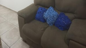 Un sofà marró amb tres coixins blaus amb textura davant d'una paret sense decoració i un paviment de rajoles.