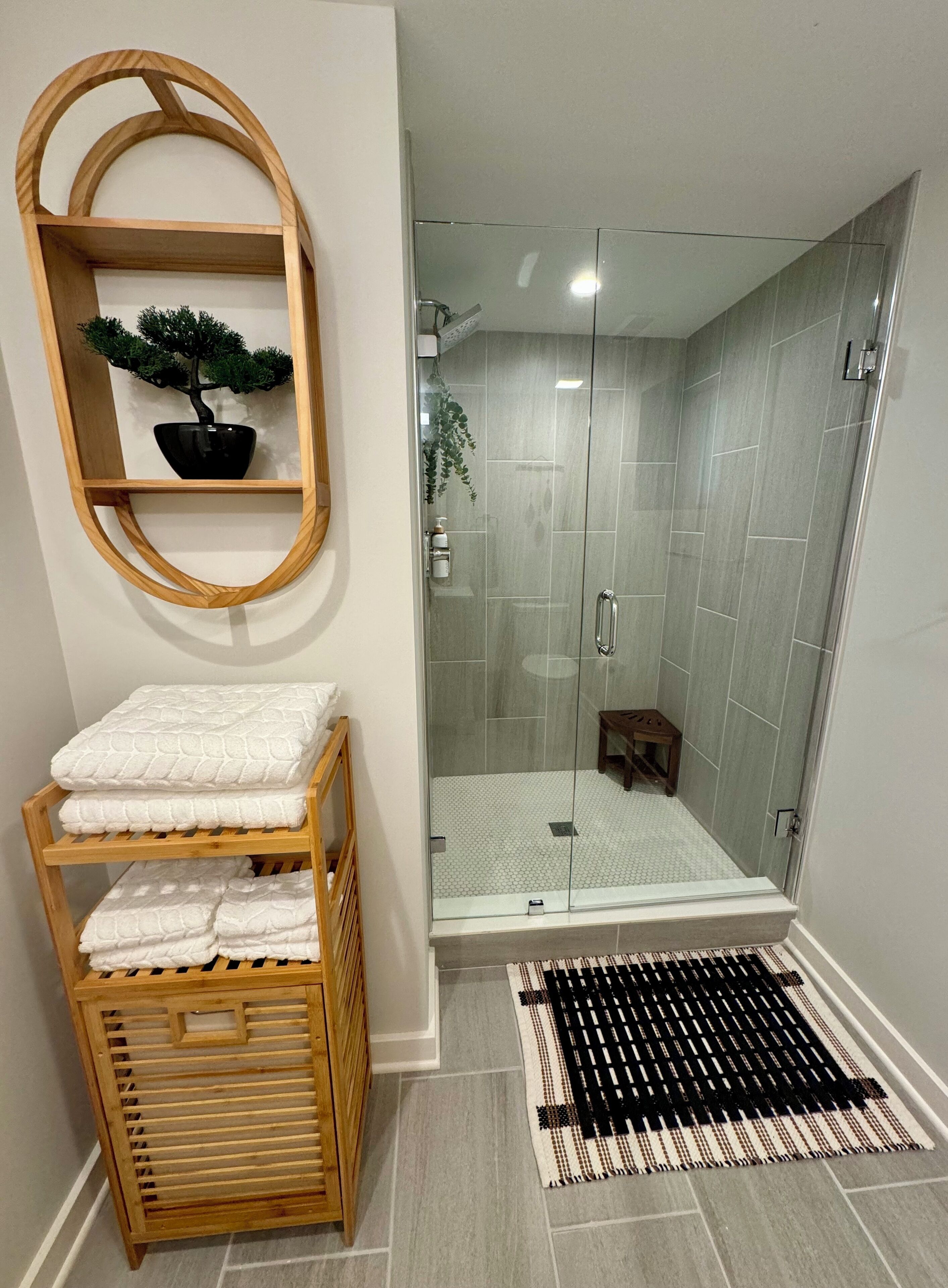 Une salle de bain moderne avec une douche à parois de verre, une étagère en bois ornée d’une plante en pot et un panier tressé.