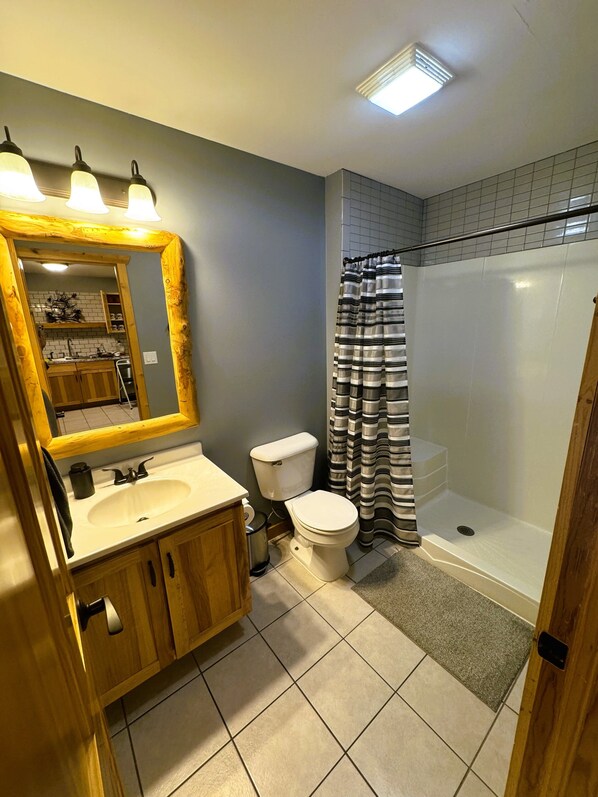 Un baño con tocador de madera, inodoro, ducha con cortina a rayas y espejo con luces sobre el lavamanos.