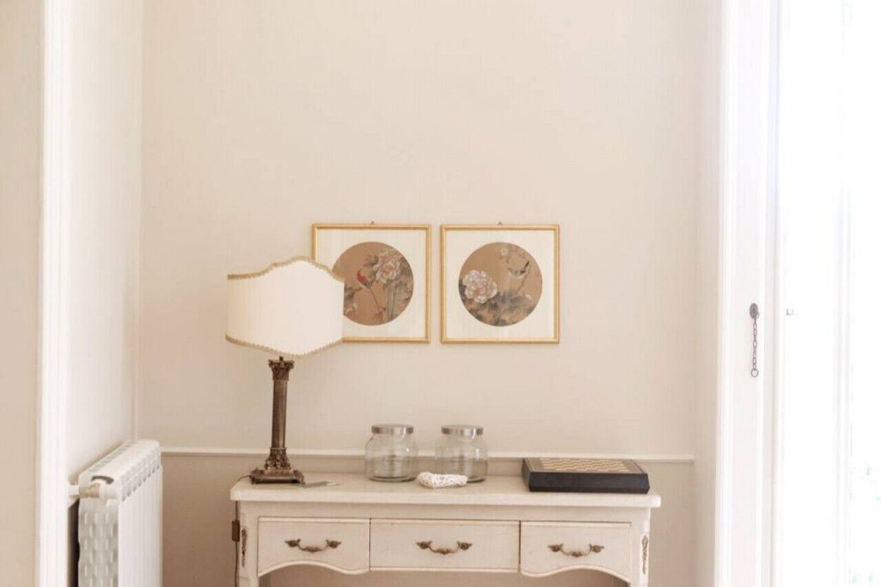A room with a white radiator, a white console table with two glass jars, a lamp, and two framed floral artworks on the wall.