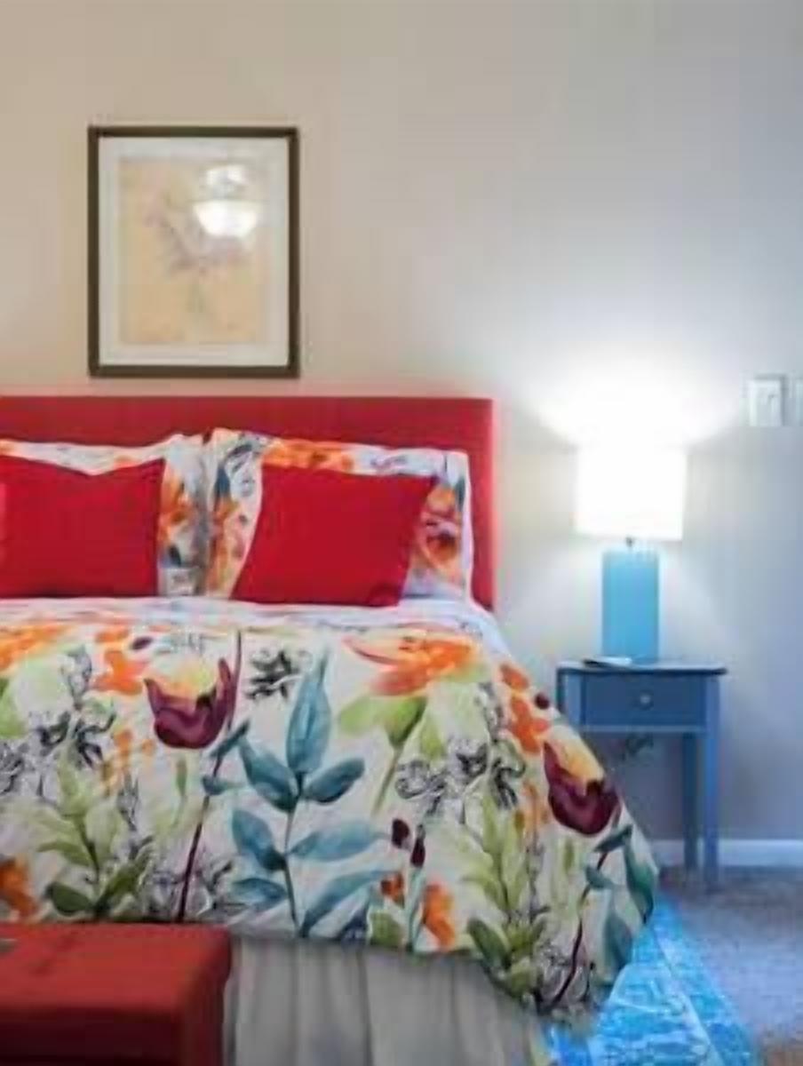 A bedroom with a bed, red pillows, a floral bedspread, a nightstand with a lamp, and a framed picture on the wall.