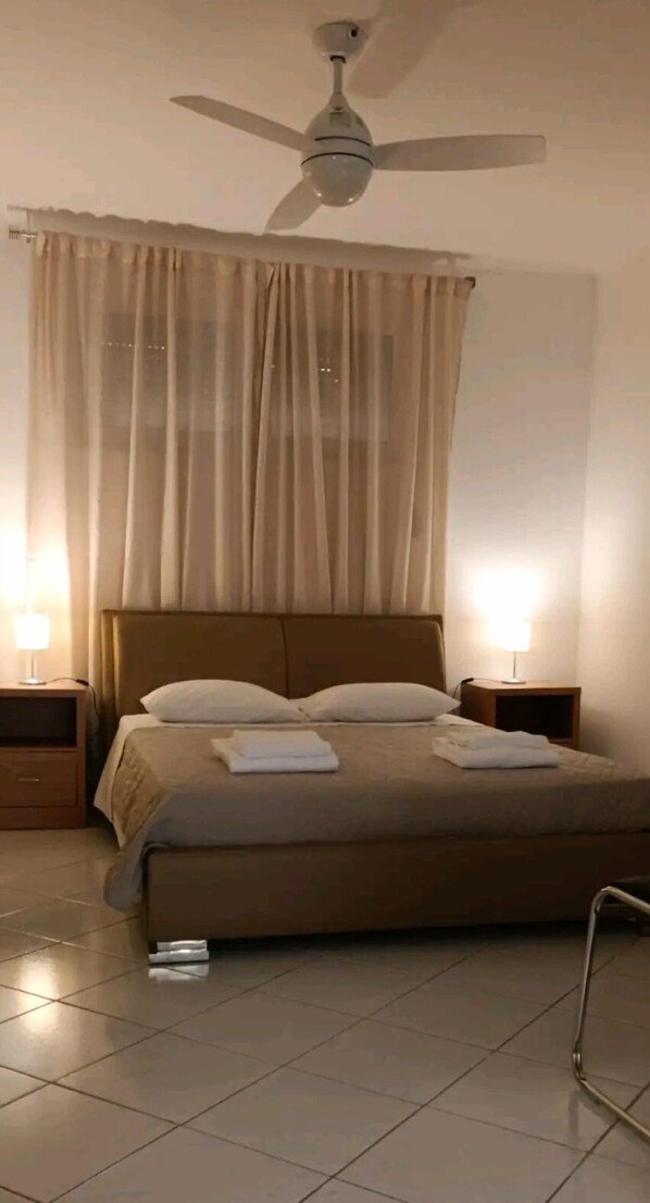 A bedroom with a bed, two bedside tables, a ceiling fan, and sheer curtains.