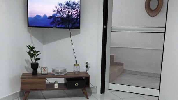 A modern living room with a flat-screen TV mounted on the wall, a wooden console table with decorative items, a woven rug, and a small plant. - Dimitri's Cosy Home in Nea Ionia with wifi,a/c, kitchen and sunny yard  (Nea Ionia)