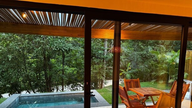 A modern outdoor pool area with a wooden table and chairs, surrounded by lush greenery.