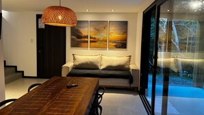A modern living room with a wooden dining table, a sofa, and a large window offering a view of trees.