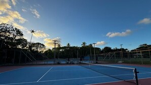 An outdoor tennis court with a net, surrounded by trees and a clear sky.
