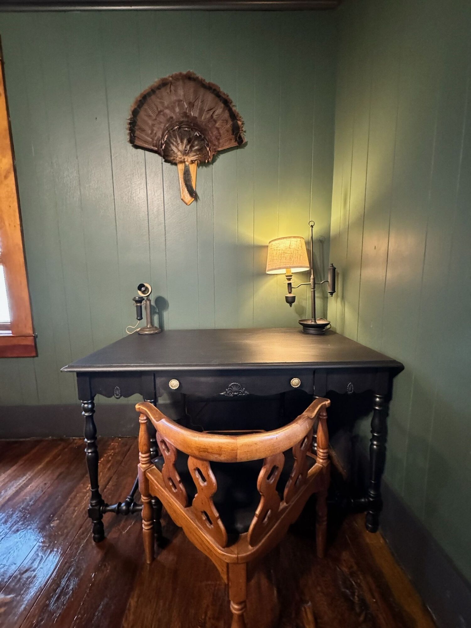 Un bureau en bois, une lampe, une chaise et un ventilateur fixé au mur.