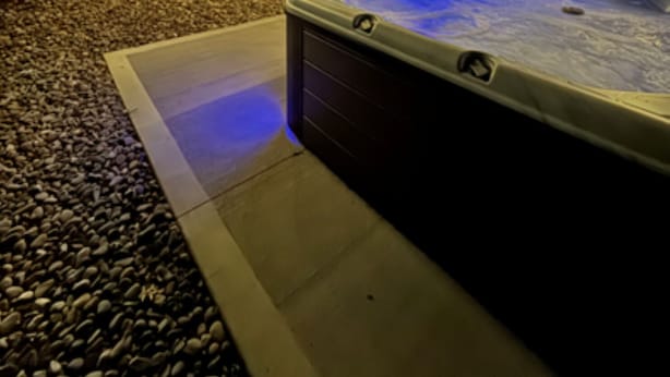A hot tub with a blue light, surrounded by a deck with string lights, and a wooden building in the background.