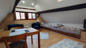 A cozy loft-style room with a wooden dining table, a blue chair, a sofa with a floral cover, and a window with wooden shutters. - Open loft apartment / parking space (Alsfeld)
