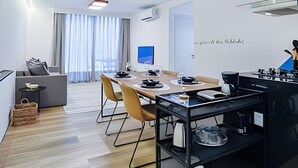 A modern, well-lit room with a dining area, a sofa, and a kitchen area with a coffee machine. - HomyCO - Conforto na esqui praia (Niteroi)