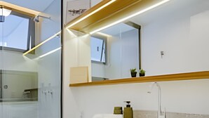 A modern bathroom with a glass shower enclosure, a white toilet, and a vanity with a large mirror and countertop. - HomyCO - Conforto na esqui praia (Niteroi)