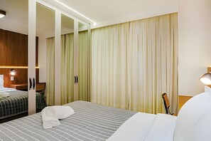 A hotel room with a bed, a seating area with a chair, a wooden cabinet, and a large window with curtains. - HomyCO - Conforto na esqui praia (Niteroi)