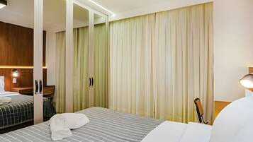 Quarto de hotel com uma cama, área de estar com uma cadeira, armário de madeira e uma janela grande com cortinas.