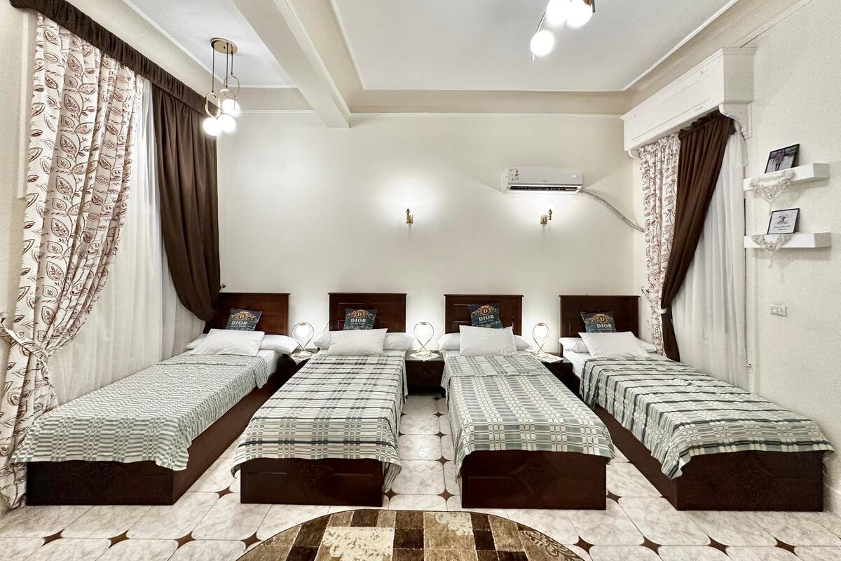 Habitación de hotel con cuatro camas individuales, cada una con cabecera y almohada con el logotipo de Dior. Encima de las camas hay un espejo grande, una lámpara de techo y un equipo de aire acondicionado en la pared.