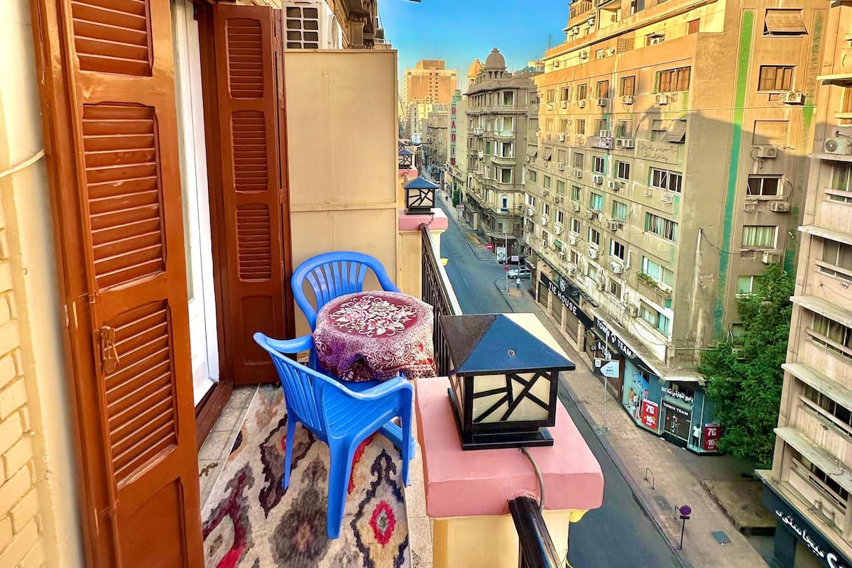 Un balcón con una silla azul y una mesa pequeña, con vista a una calle con edificios y tiendas.