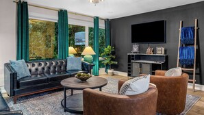 A modern living room with a black leather sofa, brown armchairs, a round coffee table, and a flat-screen TV mounted on the wall.