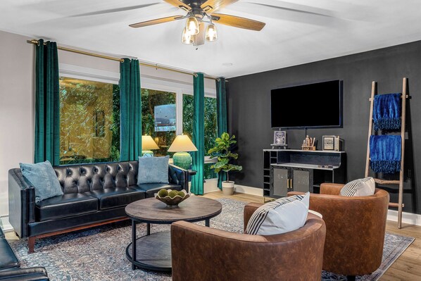 Een moderne woonkamer met een zwarte leren bank, bruine fauteuils, een ronde salontafel en een flatscreen televisie die aan de muur is bevestigd.