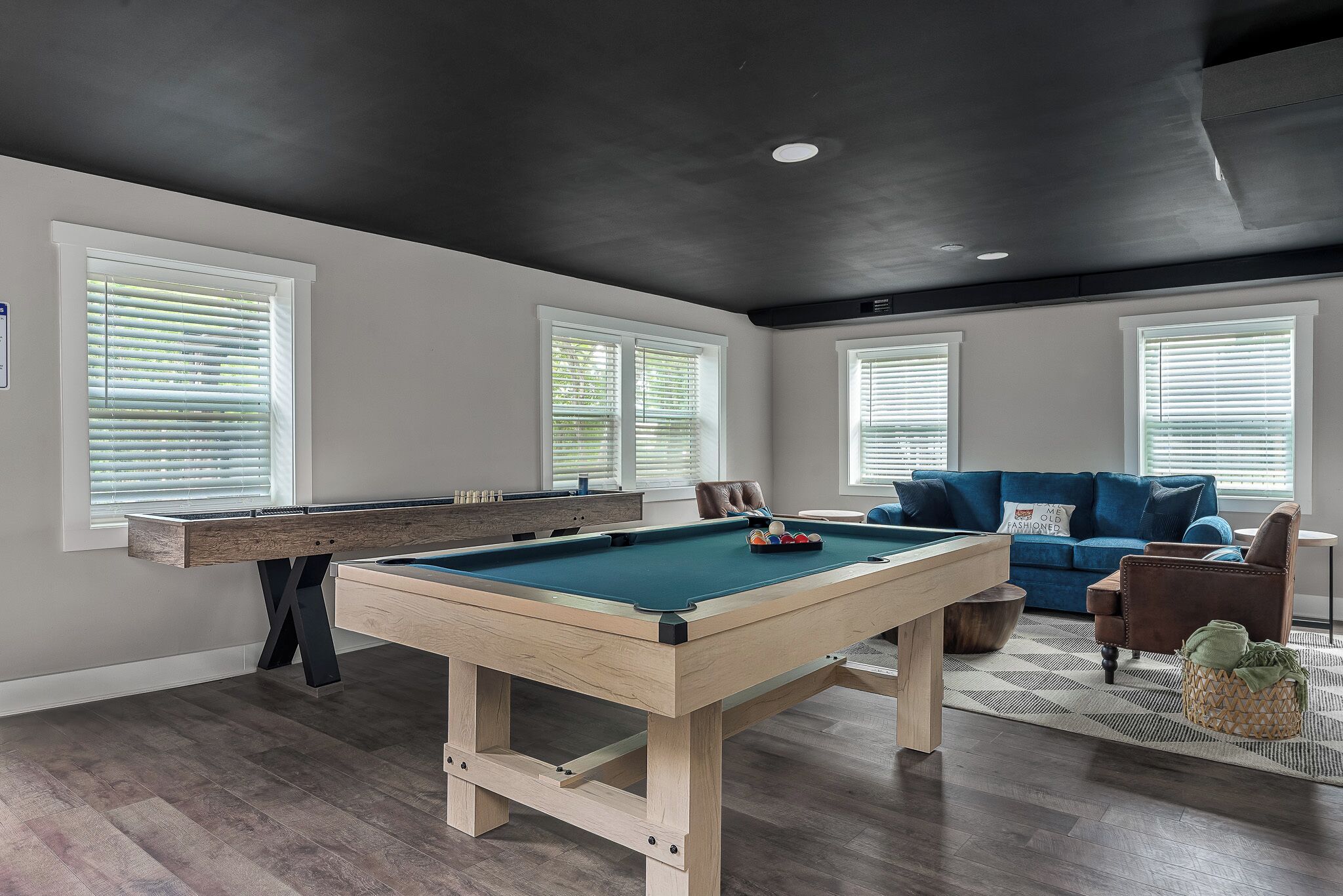 A pool table in a room with a dark ceiling, large windows, a blue sofa, and a brown leather chair.