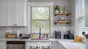 A modern kitchen with white cabinets, a built-in dishwasher, a window with blinds, and various kitchen items on the counter.