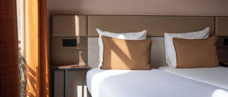 Una habitación de hotel con cama, dos almohadas, una mesita de noche y una ventana con cortinas.