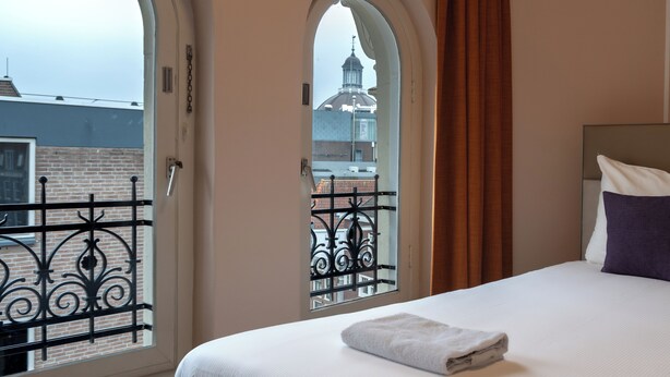 A hotel room with a bed, a window with a view of a building, and a curtain. - CS Hotel (Amsterdam)