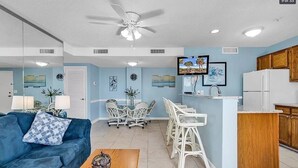 A modern living room with a blue sofa, a coffee table, and a ceiling fan. There is a kitchen area with a refrigerator, wooden cabinets, and a bar counter. - Flipflops & fun! Charming 2-bedroom ocean view condo at Myrtle Beach Resort (Myrtle Beach)