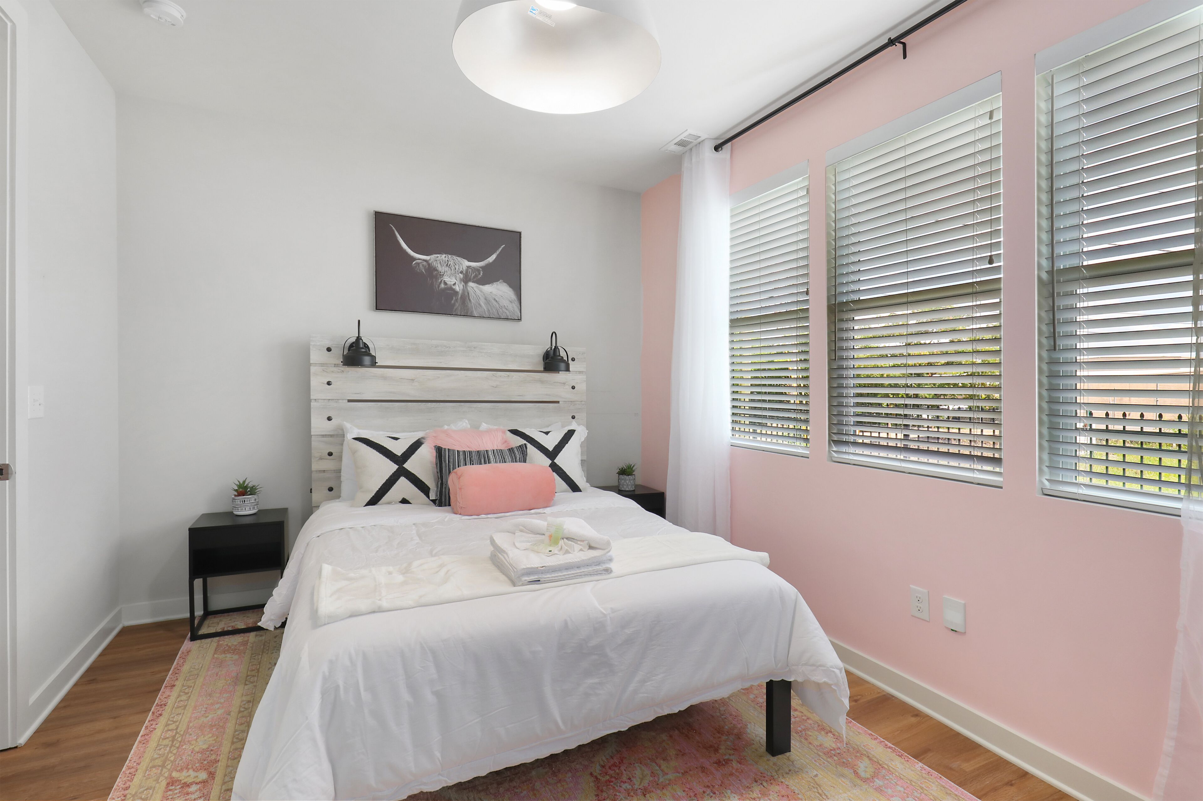 Une chambre à coucher comprenant un lit, des tables de chevet, une commode, une étagère murale, un tableau et une fenêtre avec des stores.