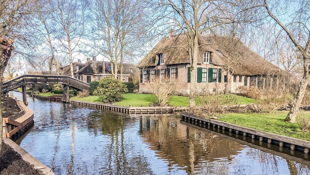 Un canal con un puente, una casa de techo de paja y una zona cubierta de césped.