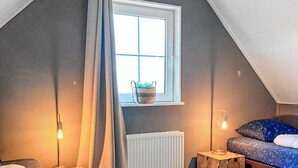 Un dormitorio acogedor en el ático, con techo inclinado, una ventana con vistas, un radiador, una cama con colcha estampada y dos lámparas de noche.