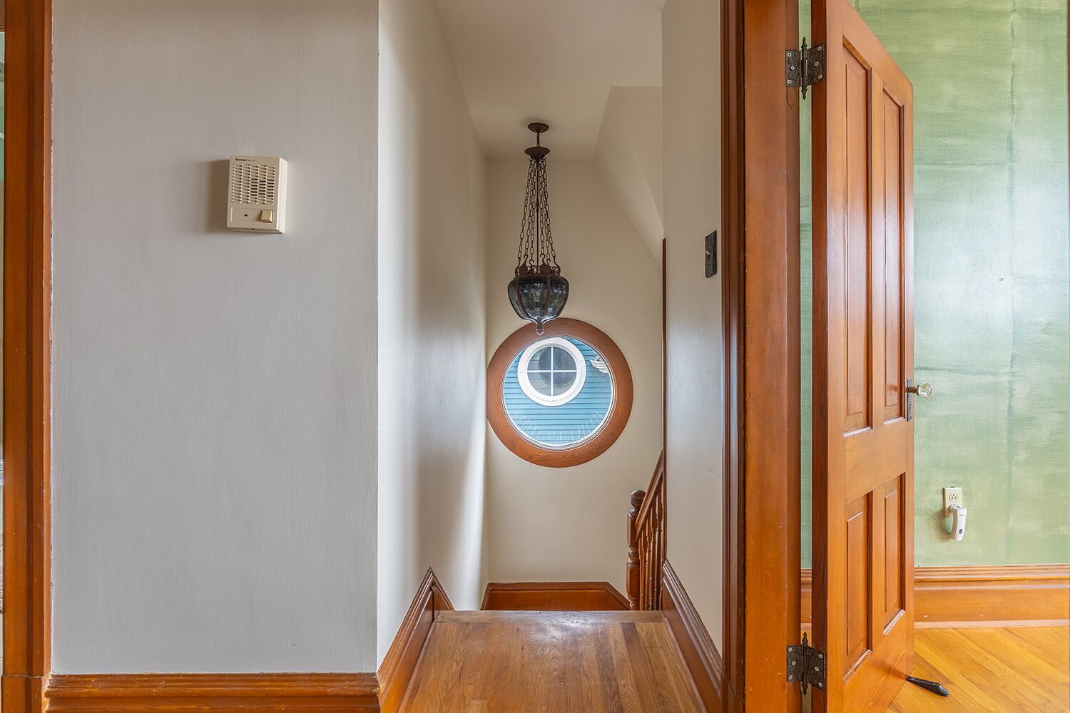 A hallway with a wooden door, a round window with a blue frame, and a hanging light fixture.