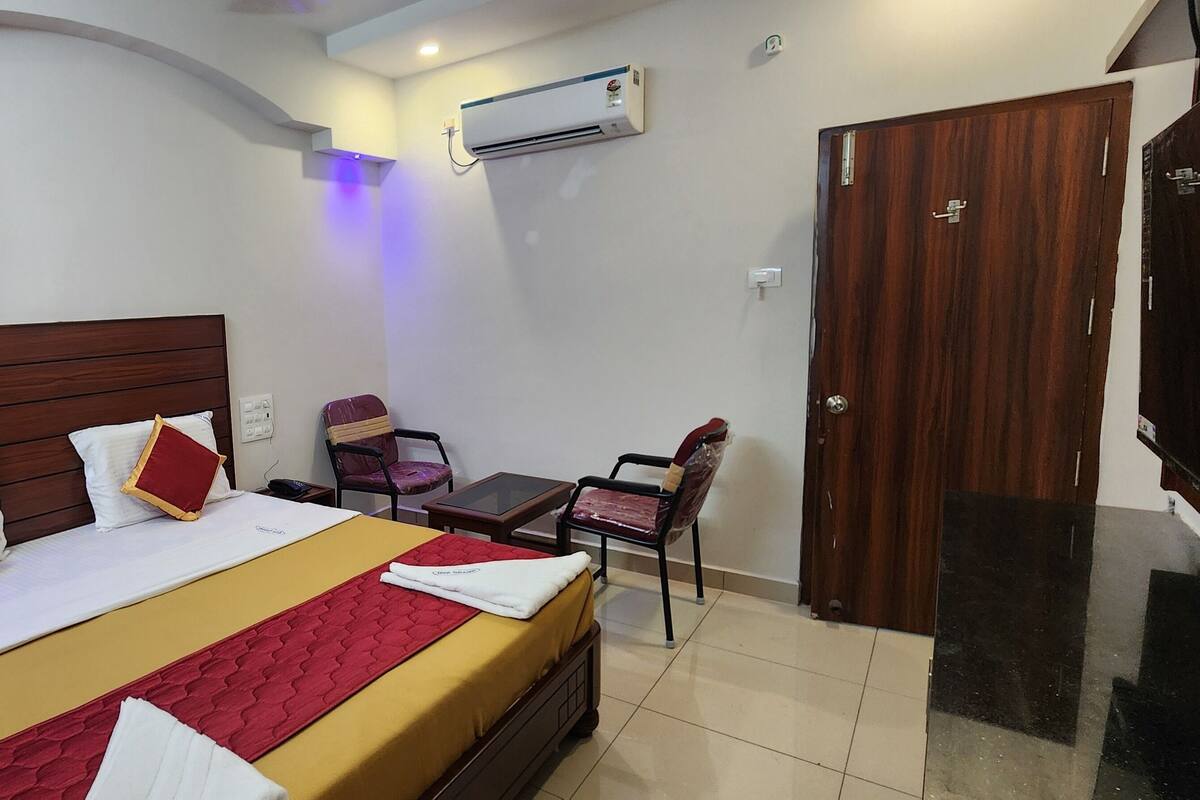 Hotellihuone, jossa on puinen sänky, pieni pöytä, tuoli, ilmastointilaite ja puinen ovi.