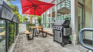 Une terrasse extérieure avec un barbecue, une table en bois, un banc et un parasol rouge.