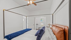 Un lit à baldaquin avec une couverture bleue et deux oreillers, un ventilateur de plafond et une vue sur un couloir avec une porte.