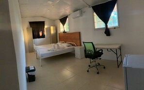 Ein Zimmer mit einem Einzelbett, einem Holzkopfteil, einem Schreibtisch mit Stuhl, einem kleinen Nachttisch und einem Fenster mit Vorhängen.