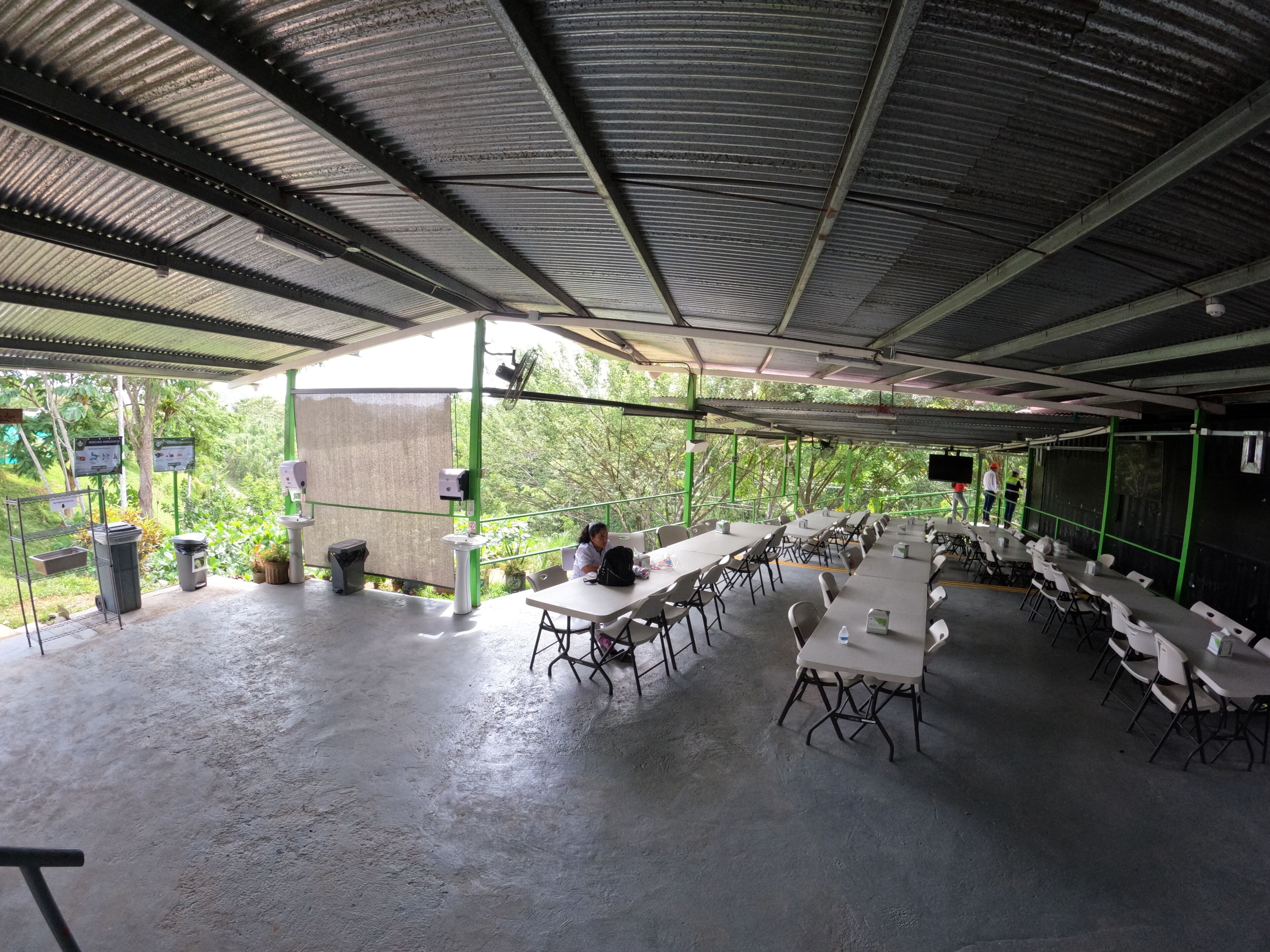 An outdoor covered dining area with tables and chairs, a trash bin, and a green railing.