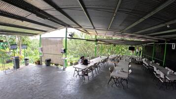 An outdoor covered dining area with tables and chairs, a trash bin, and a green railing.