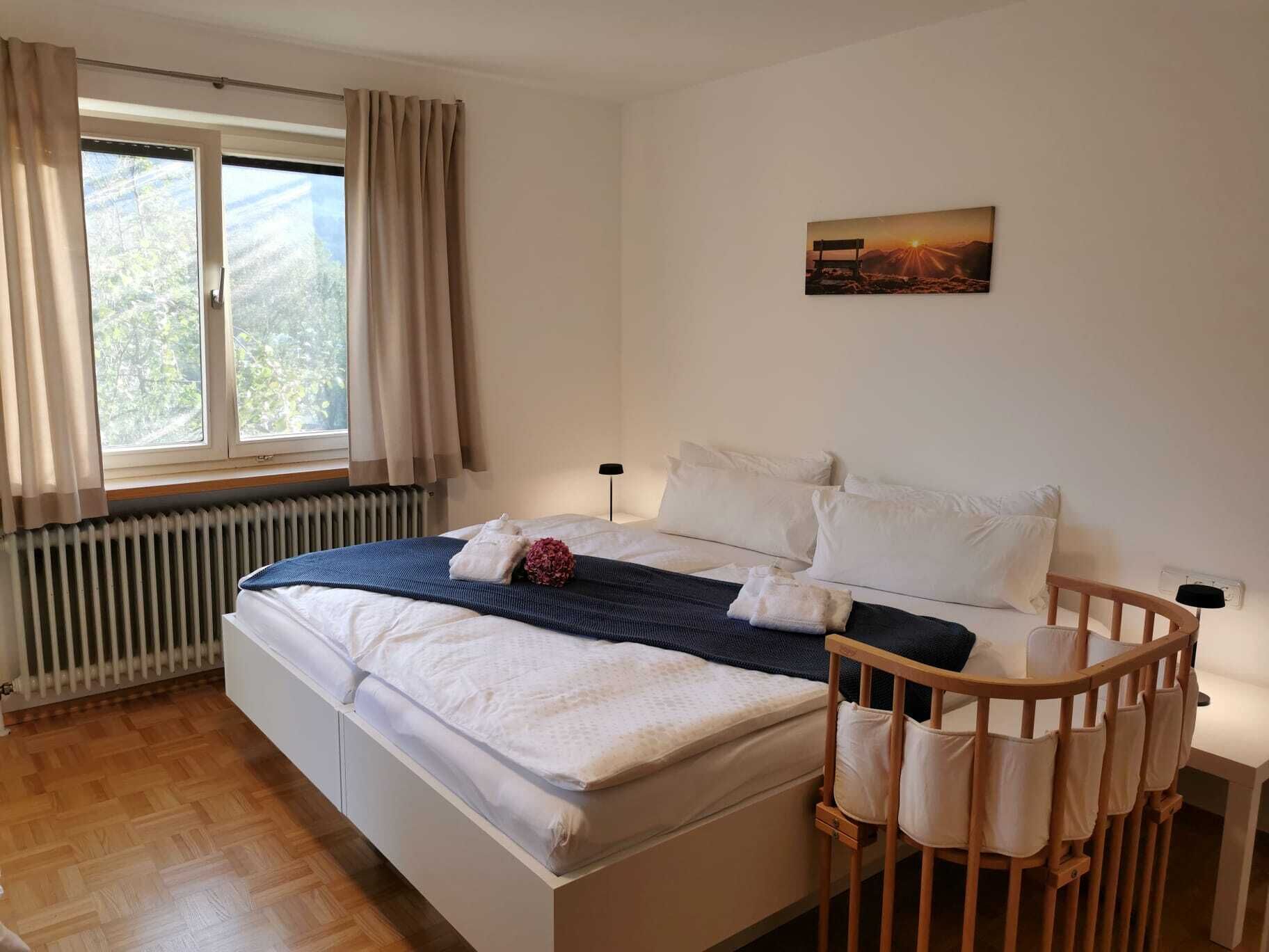 A neatly made bed with white linens, a blue blanket, and a pillow with a red flower. There is a wooden crib with white cushions next to the bed. A painting of a sunset is on the wall above the bed.